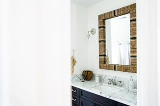 Bathroom design similar to a beach home with a woen mirror, marble countertops, dark blue drawers.