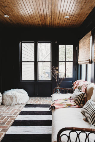 Side view of a sunroom with walls painted black, wood panels on the ceiling, brick floors, and a stiped area rug.