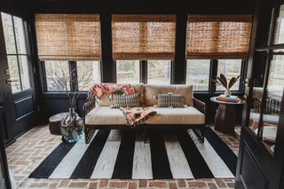 A sunroom with black wall paint, striped rug, rustic couch and blinds.