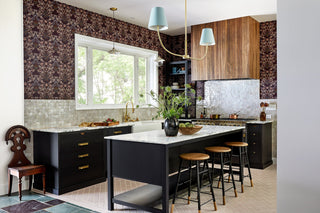 Wideshot of a remodeled kitchen with dark-colored custom cabinets, wallpaper for the kitchen, white tiles against the range, wood hood over the oven.