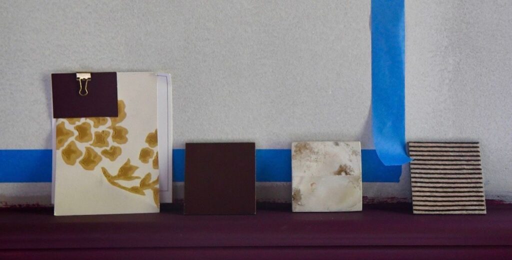 Fabric, paint, and stone material swatches — deep brown, striped textile, and marbled stone — arranged on a mantel to test a cohesive timeless color palette for interior design.