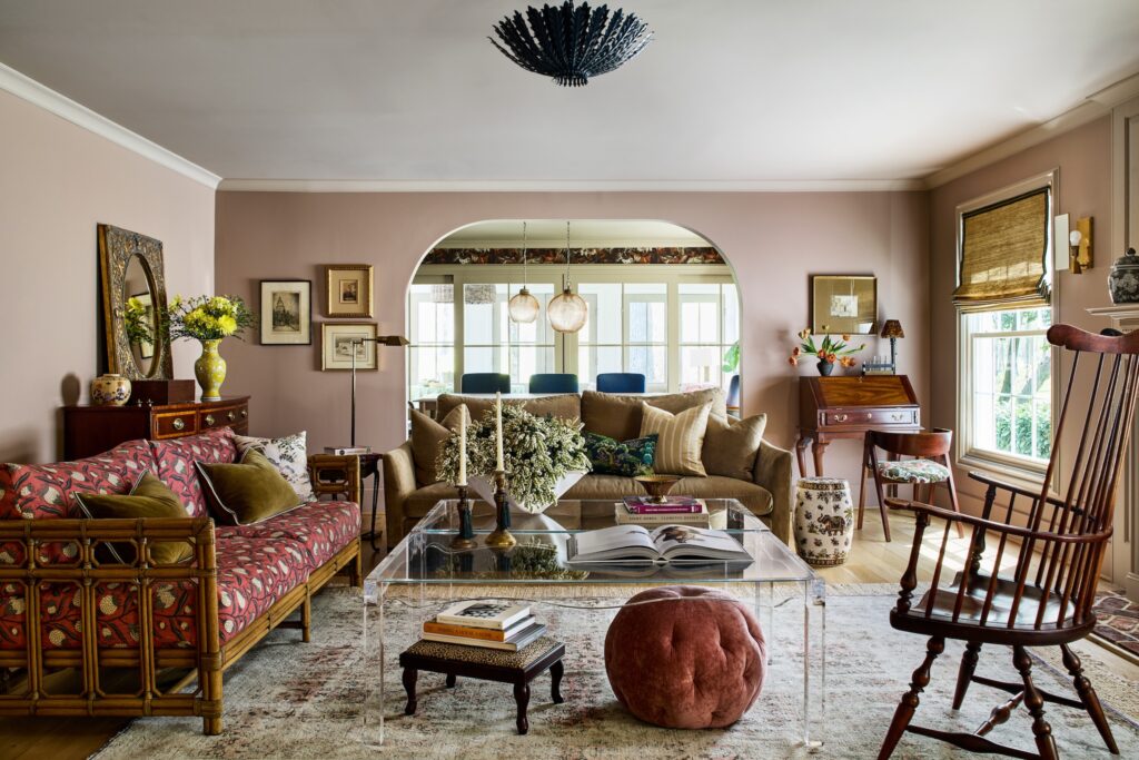Eclectic traditional living room with blush pink walls, rattan sofa in a red patterned fabric, tan sofa with layered throw pillows, clear acrylic coffee table styled with books and flowers, vintage wood rocking chair, and arched doorway leading to a bright dining room with large windows.
