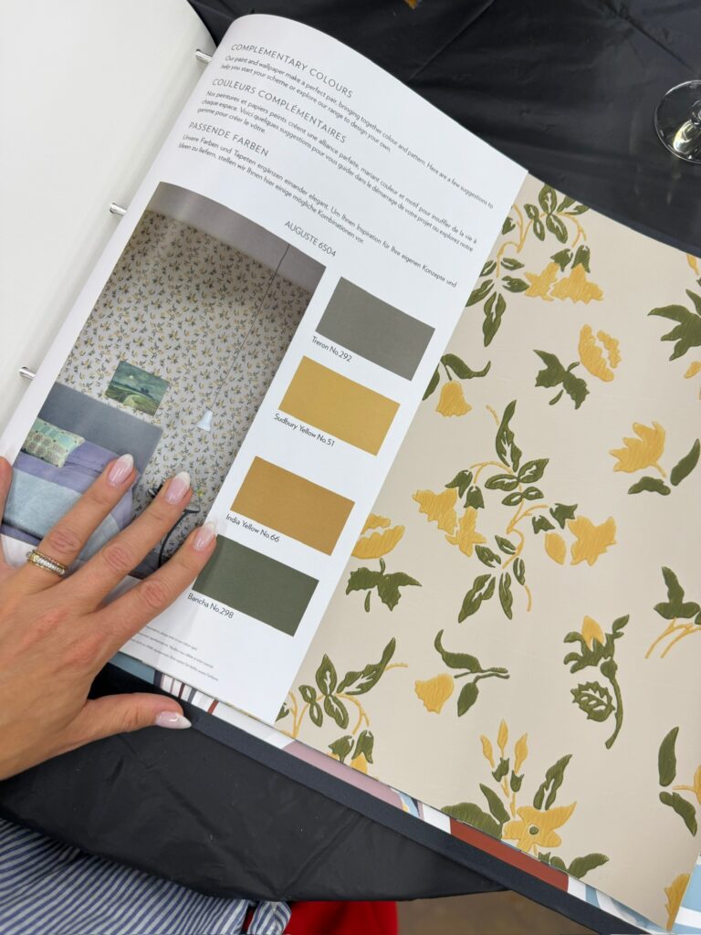 Designer flips through a wallpaper sample book showing a floral yellow and green print paired with complementary paint chips, a key step in building a timeless whole-home color scheme.